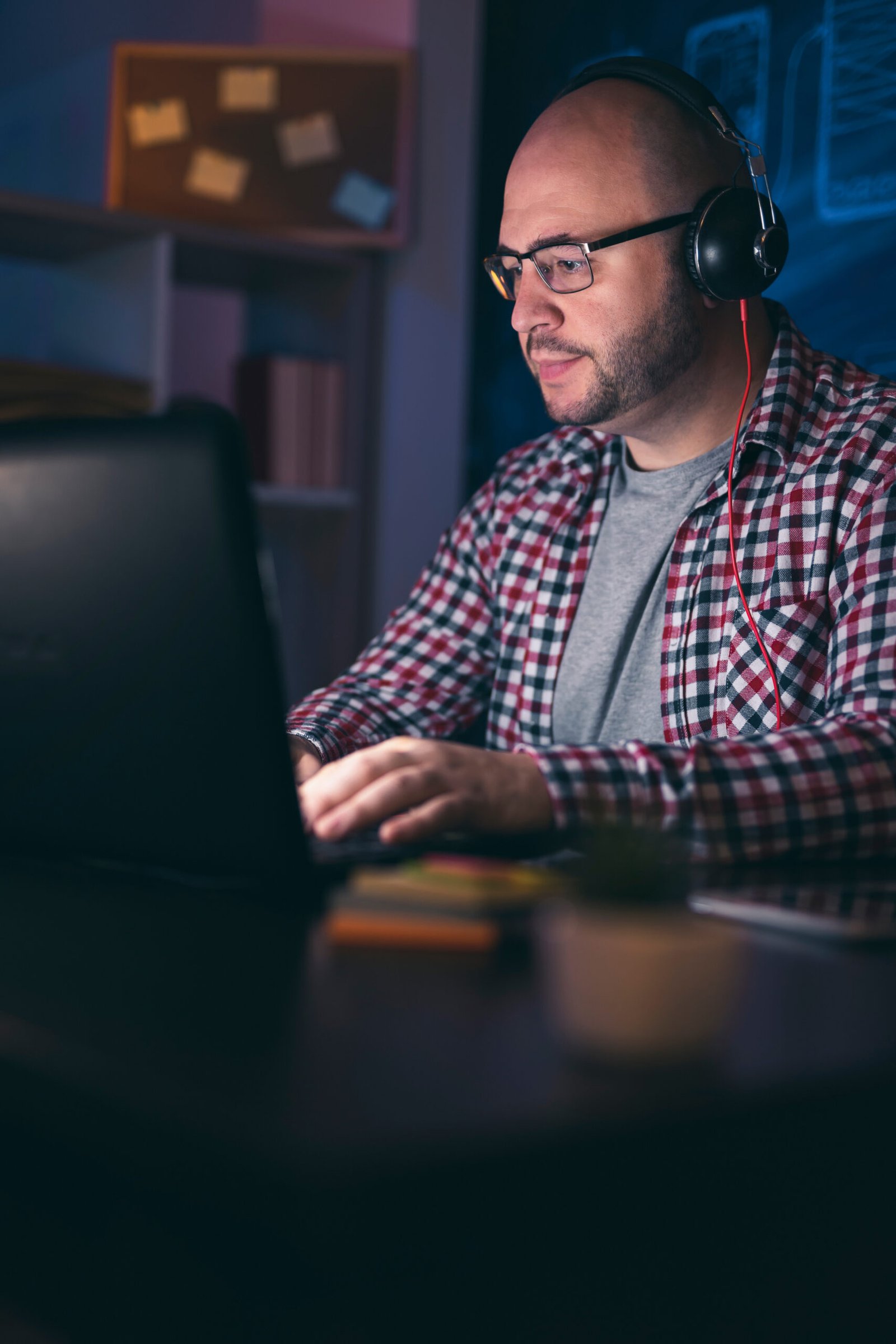 Man wearing headset working on laptop computer in home office late at night; web developer working overtime